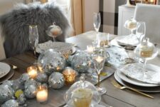 a shiny holiday tablescape with silver disco balls forming a table runner, candles, white porcelain and gold cutlery