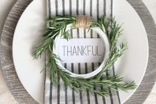 a simple and modern Thanksgiving place setting with a plywood placemat, a white plate, a striped napkin and a greenery wreath