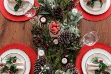 a stylish Christmas tablescape with evergreens, pinecones, candles, red placemats, white porcelain and napkins accented with greenery