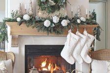 a chic Christmas mantel with an evergreen and white flower garland, candles, a mirror with a greenery and whie bloom wreath plus white stockings