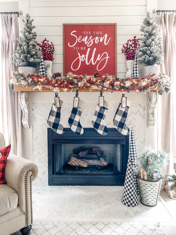 a cozy farmhouse christmas mantel with a snowy pinecone and light garland, red berries, snowy mini trees and buffalo check stockings