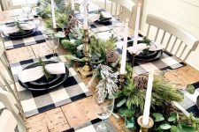 a farmhouse Christmas table with an evergreen and pinecone runner, tall and thin candles, buffalo check placemats, black plates