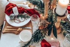 a lovely Christmas tablescape with greenery, pinecones, wooden candleholders with pillar candles, red napkins, cutout wooden boards and bells