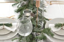 a simple modern Christmas tablescape with an evergreen runner, snowy pinecones, tall and thin candles, neutral porcelain and linens