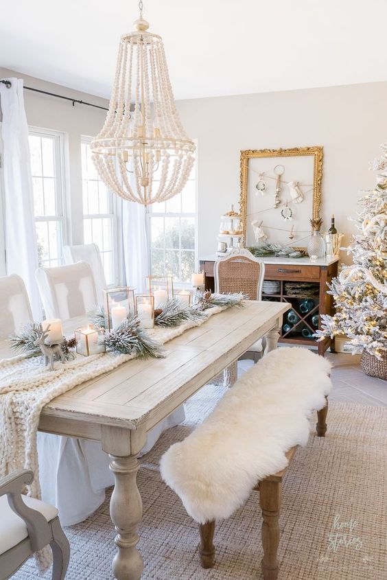 a white Christmas tablescape with a knit table runner, flocked greenery, candle lanterns and deer and a beaded chandelier over the table
