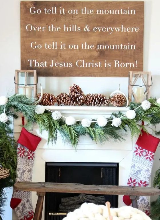 an extremely cozy Christmas mantel with snowy pinecones, candle lanterns, a white pompom garland and pinted stockings, a large sign