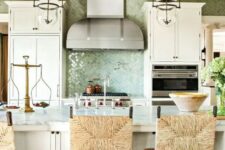 a beautiful coastal kitchen with white vintage cabinets, a large kitchen island with wicker chairs, glass pendant lamps and a green tile backsplash
