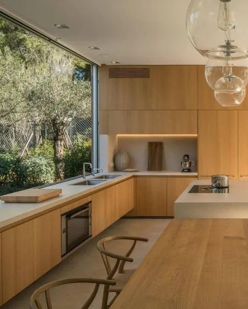 A beautiful contemporary kitchen with sleek light stained cabinets, white stone countertops, built in lights and a glazed wall with garden views