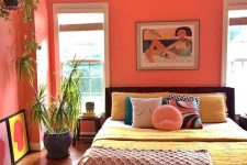 a bold bedroom with coral walls, a dark bed with colorful bedding, a tassel blanket, colorful artworks and potted plants