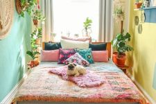 a bright boho bedroom with a mint green and yellow wall, a stained bed with colorful bedding, a bright rug and pillows and potted plants