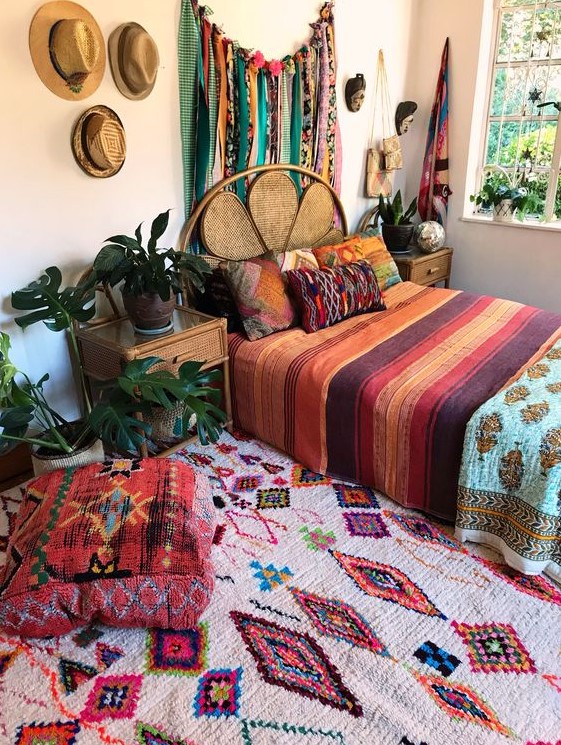 a bright boho bedroom with a rattan bed and wooden nightstands, a bold rug and bedding, a bunting and hats for decor on the wall