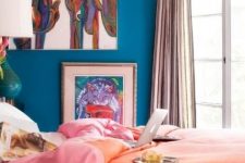 a colorful maximalist bedroom with blue walls, bright artworks, colorful bedding and curtains plus a green table lamp
