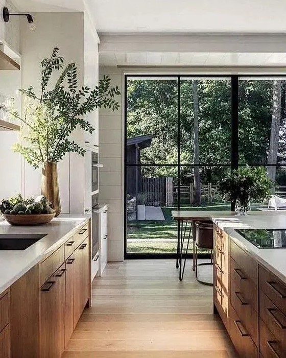 A contemporary kitchen with light stained cabinets, white stone countertops, a dining space in front of the glazed wall that gives garden views