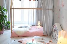 a delicate pastel girl’s room with grey walls, a house-shaped bed with pink bedding and a grey canopy, a grey rug and a playhouse