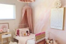 a dreamy pink girl’s bedroom with a watercolor yellow and hot pink accent wall, a white metal bed with pink bedding, a pink tassel chandelier