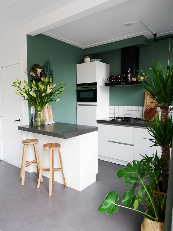 a green kitchen with white tiles, grey stone countertops, a black hood and potted plants and blooms