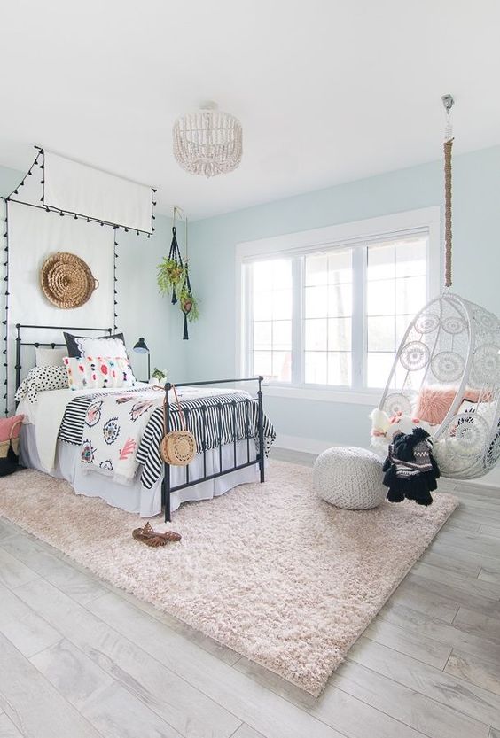 a lovely coastal girl bedroom with light blue walls, a black metal bed with a canopy, a pendant chair and a fluffy rug