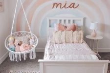 a lovely girl’s room with a rainbow accent wall, a white bed and a nightstand, pendant chair, a pink printed rug is a cute and cozy space