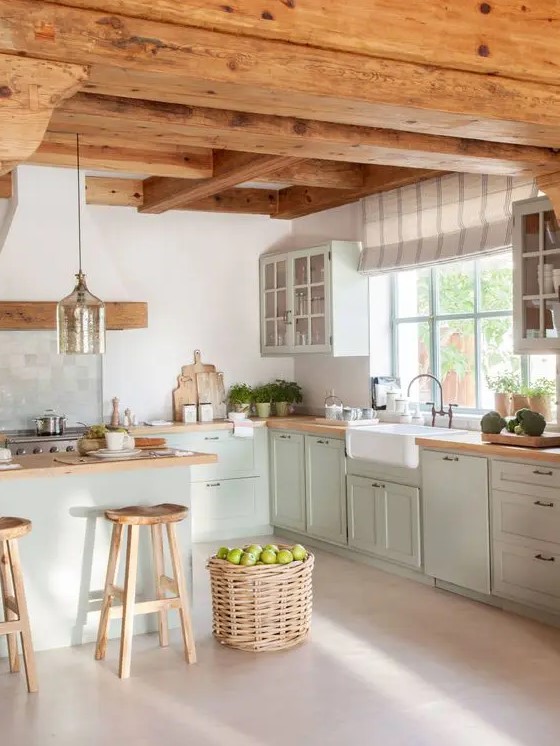 a modern cottage kitchen with wooden beams on the ceiling, light green shaker style cabinets, butcherblock countertops, a basket for storage and a pendant lamp