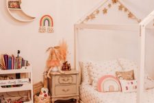 a neutral boho girl bedroom with a wooden house-shaped bed with neutral bedding, a storage unit, a vintage nightstand and a moon-shaped shelf