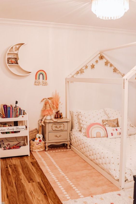 A neutral boho girl bedroom with a wooden house shaped bed with neutral bedding, a storage unit, a vintage nightstand and a moon shaped shelf