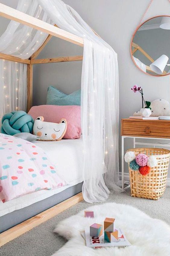 A neutral girl's room with a house shaped bed with pastel bedding, a nightstand, a basket with pompoms and layered rugs