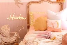 a pink and blush girl’s room with color block walls, a blush bed, blush bedding, mellow yellow pillows, a tassel chandelier and a wicker baby carriage