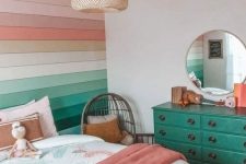 a pretty rainbow girl bedroom with a rainbow accent wall, a bed with pastel and neutral bedding, an emerald dresser with a round mirror