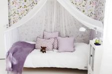 a romantic girl’s room with floral wallpaper, a white bed with lilac and white bedding, a white canopy, a white cabinet and a purple printed rug