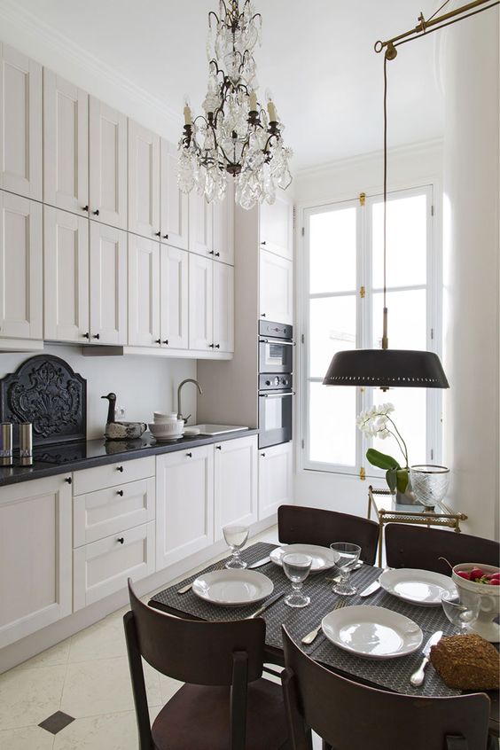 a small and chic French style kitchen with black coutnertops, a crystal chandeliers, a small dining zone with an additional lamp