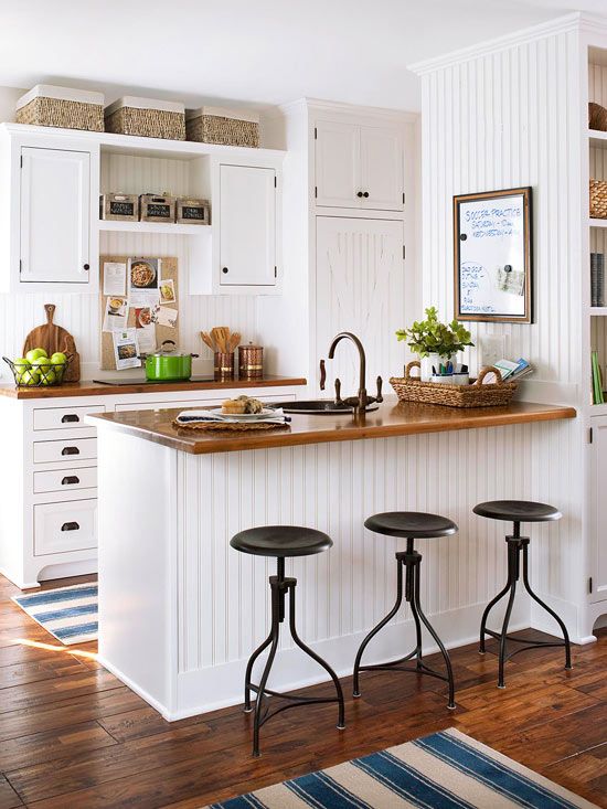 a small white farmhouse kitchen with stained wooden countertops, black stools, boxes and baskets is welcoming