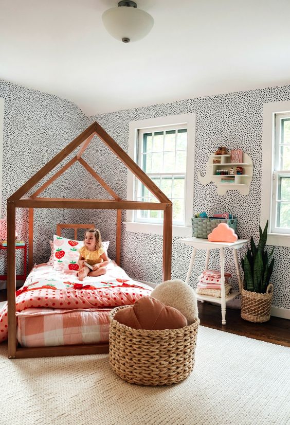 A whimsy girl's room with spotted wallpaper, a house shaped bed with bright bedding, a white nightstand, a basket with pillows