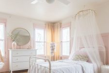 an airy girl’s bedroom with pink paneling, a white bed with neutral bedding and a canopy, a white dresser