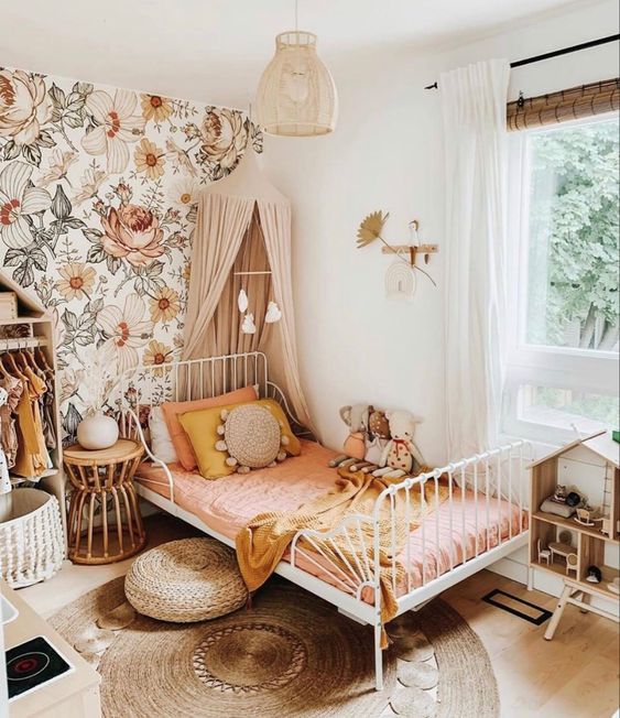 An earthy tone girl's bedroom with floral wallpaper, a white metal bed with pink bedding, a canopy, an open wardrobe and a play house