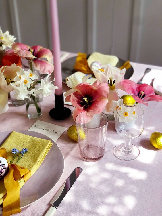 a beautiful and bright Easter table with a pink tablecloth, candles and blooms, yellow napkins, gilded egs and some white flowers is amazing