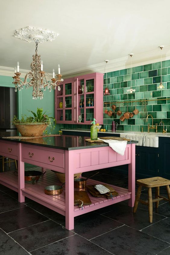 a bold kitchen with black lower cabinets, upper pink ones, a pink kitchen island, a bold green tile backsplash and a chic chandelier