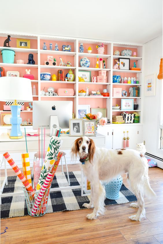 a bright and welcoming home office wiht a large white storage unit with salmon pink backing, a white desk and a plaid rug, colorful accessories and a lamp