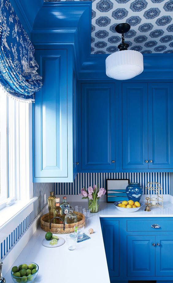 a bright blue kitchen with a striped backsplash and a bold wallpaper ceiling looks siper bright and very elegant, seaside-inspired