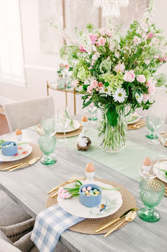 a fresh Easter tablescape with pink and white blooms plus greenery, woven placemats, pastel egg candles and checked napkins, green glasses