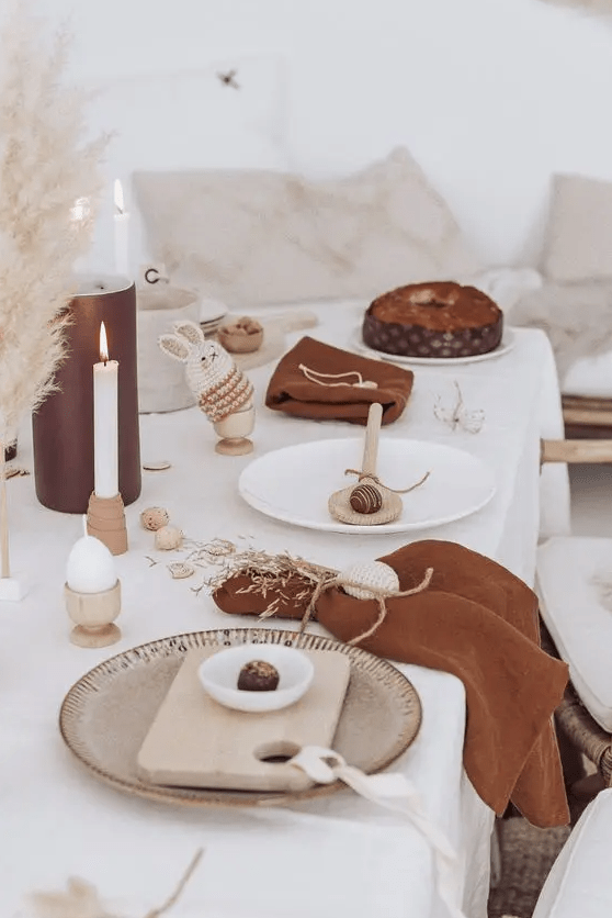 a fun modern Easter tablescape with a white tablecloth and rust-colored napkins, candles, pampas grass, wooden boards and spoons, bunny cozies for eggs