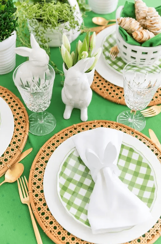a green and white Easter tablescape with a green tablecloth, rattan placemats, green plaid plates, white tulips, bunny figurines and gold cutlery