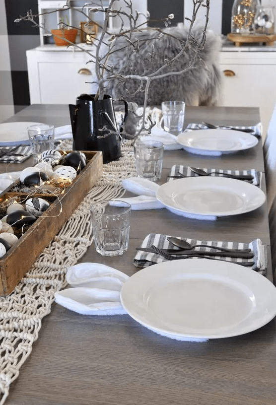 a monochromatic Easter tablescape with a macrame runner, a wooden box with dyed eggs and lights, striped napkins and black cutlery