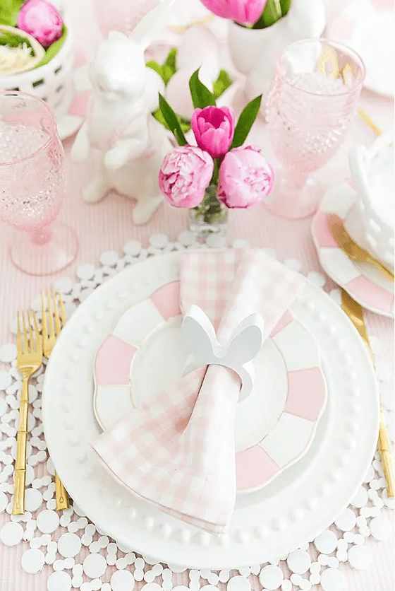 a pink and white Easter table setting with plaid plates and napkins, a cool placemat, pink glasses, blooms and porcelain bunnies