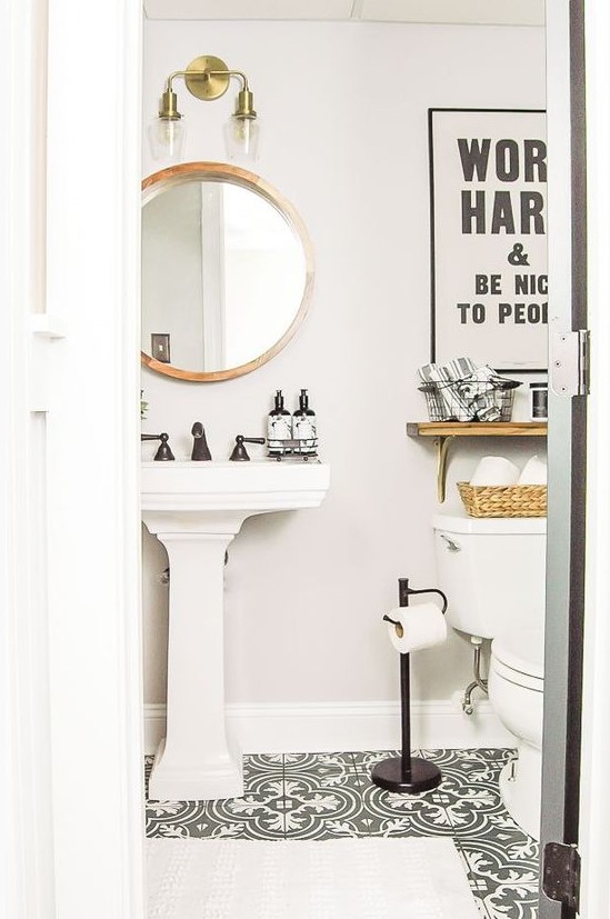 a pretty small powder room with black and white tiles, a pedestal sink, a round mirror in a wooden frame, a shelf and black fixtures