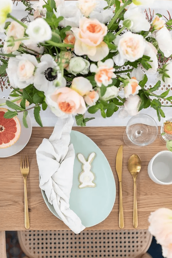 An Easter table setting with pastel blooms, a mint colored egg shaped plate, gold cutlery and bunny cookies