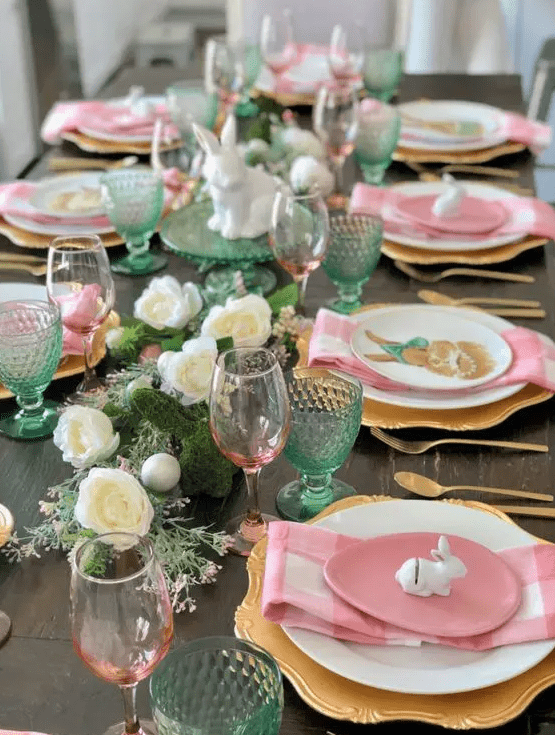 An Easter tablescape with white blooms and greenery, pink and green glasses, gold chargers and pink egg shaped plates and pink plaid napkins