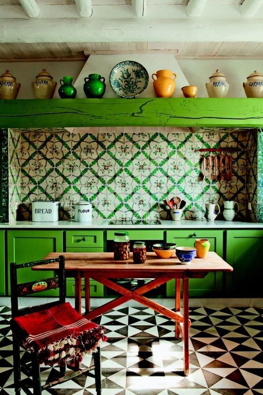 an eclectic kitchen with bold green cabinets, bright printed tiles on the backsplash, a wooden table and a chair plus colorful tableware