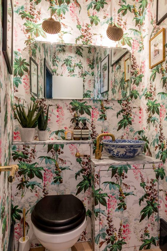 a crazy guest toilet done with bright floral wallpaper, a vanity covered with it, a mirror, some lamps and gold touches