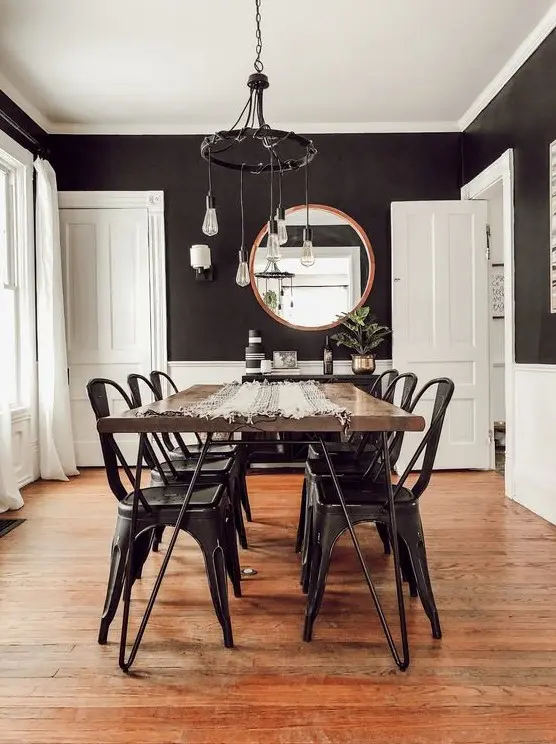 a farmhouse dining room with black walls and white paneling, a hairpin leg table, black metal chairs, a black chandelier with bulbs
