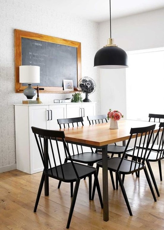 a laconic Scandinavian dining space with a chalkboard, cabinets, a stained table and black chairs, a black pendant lamp